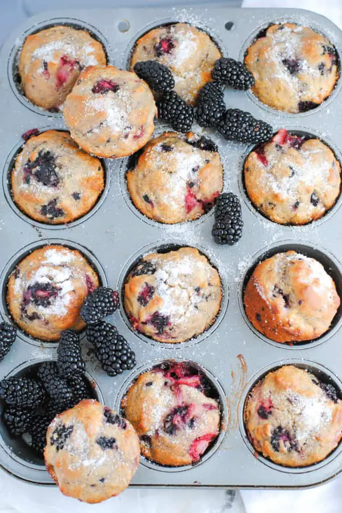 mixed berry muffins in muffin tin with blackberries around muffins which are topped with sugar