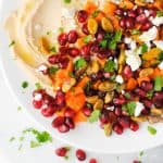 hummus topped with roasted sweet potato, pomegranate seeds, feta, pistachios, mint and parsley with a drizzle of olive oil on a white plate