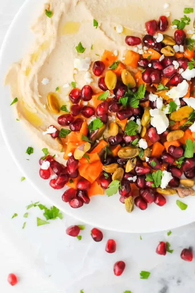 hummus topped with roasted sweet potato, pomegranate seeds, feta, pistachios, mint and parsley with a drizzle of olive oil on a white plate