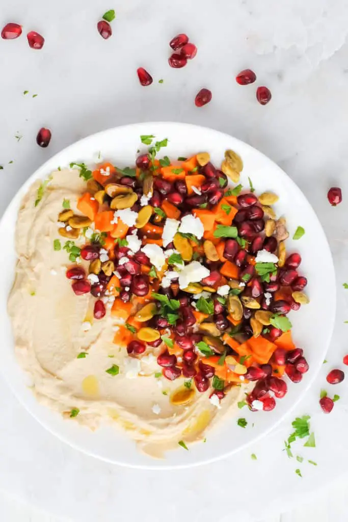 hummus with roasted sweet potato, pomegranate seeds, pistachios, feta, mint and parsley on a white plate