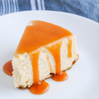 slice of cheesecake with salted caramel sauce drizzled on top set on a white plate with gray background