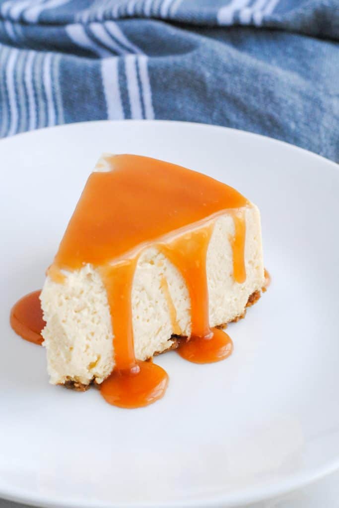 slice of cheesecake with salted caramel sauce drizzled on top set on a white plate with gray background