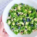 hand holding a white bowl with kale salad topped with blueberries, avocado, sliced almonds and honey lime vinaigrette dressing