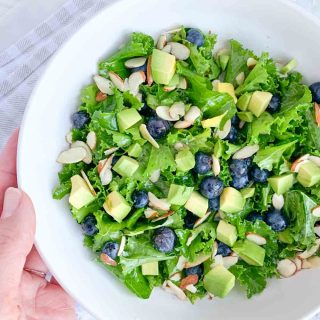 hand holding a white bowl with kale salad topped with blueberries, avocado, sliced almonds and honey lime vinaigrette dressing