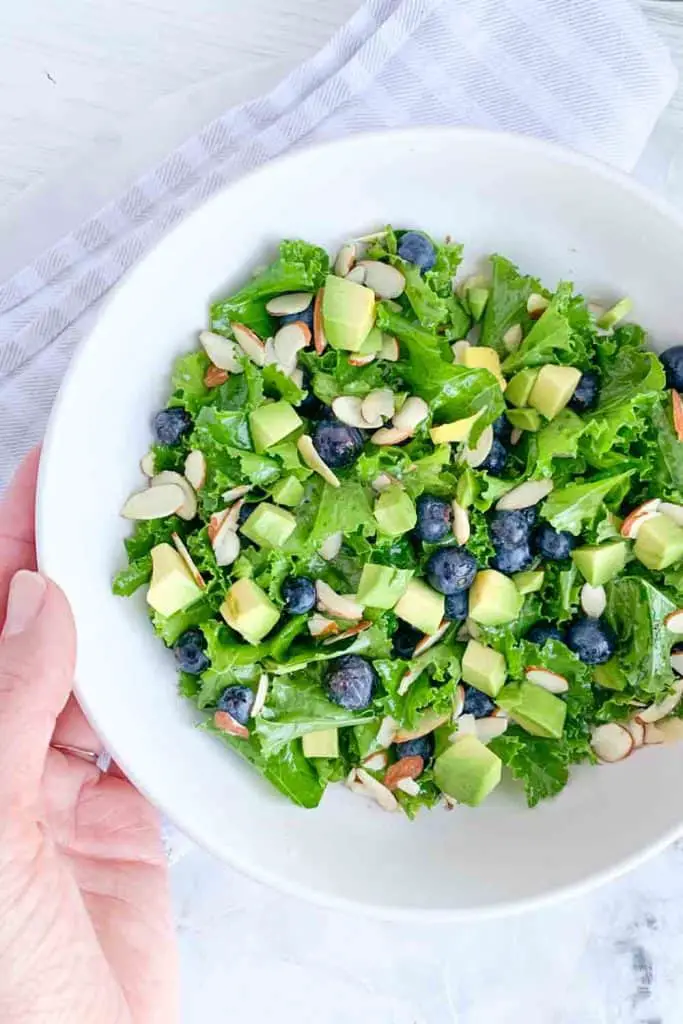 hand holding a white bowl with kale salad topped with blueberries, avocado, sliced almonds and honey lime vinaigrette dressing