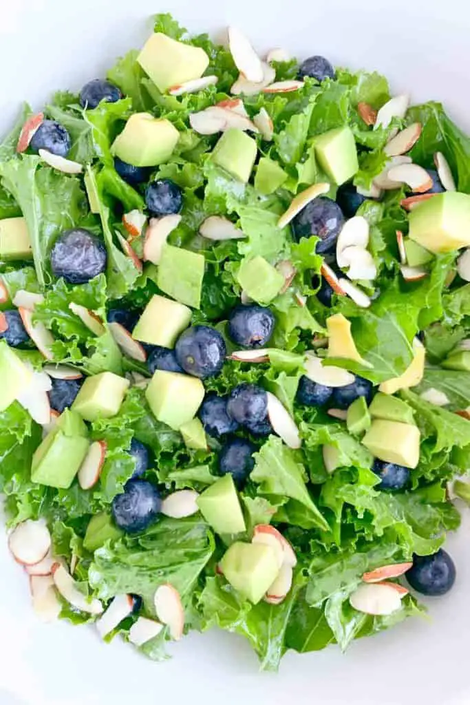 white bowl with kale salad topped with blueberries, avocado, sliced almonds and honey lime vinaigrette dressing