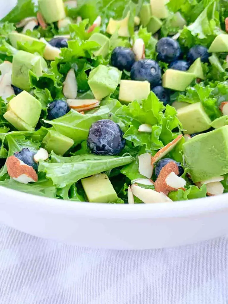 close up of white bowl with kale salad topped with blueberries, avocado, sliced almonds and honey lime vinaigrette dressing