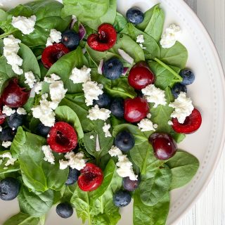 white bowl filled with spinach salad, and topped with cherries, blueberries, goat cheese, and red onion